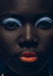 Close-up beauty portrait of Black model Awek Gak featuring bold pastel blue eyeshadow and vibrant orange lipstick in low-key lighting.