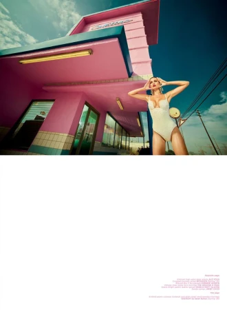 Model Karoline Niek in a white one-piece bodysuit posing in front of a retro pink building under a bright blue sky.