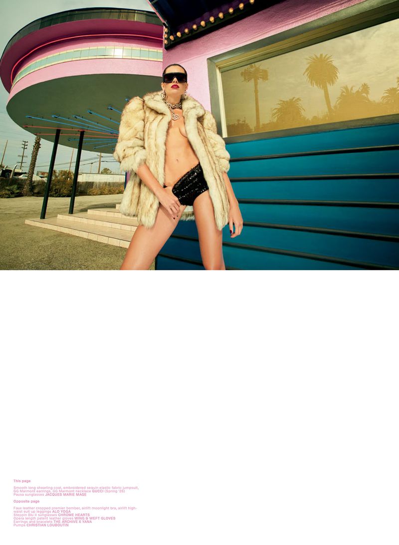 Fashion model posing outdoors in an open fur coat, black sequined briefs, and dark sunglasses beside a retro pink building.