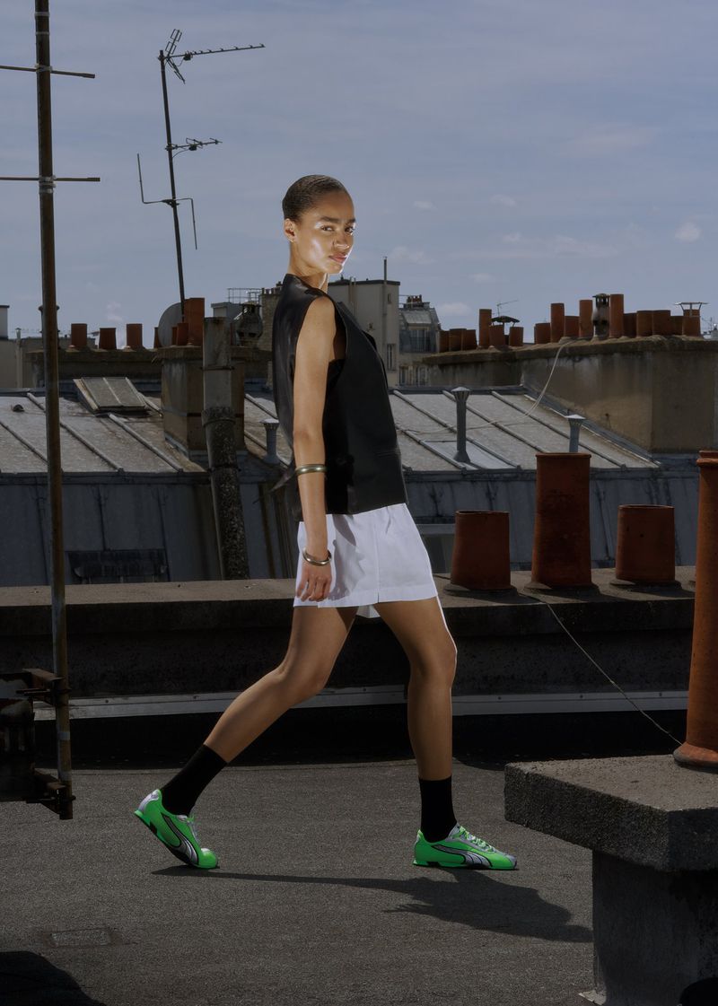 A slender person with short hair is walking on a rooftop wearing a sleeveless top, short skirt, green sneakers, with a city background.