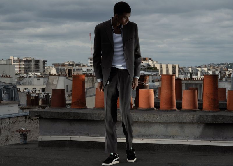 A young man in a grey suit stands on a rooftop in an urban landscape with chimney pipes and buildings under an overcast sky.