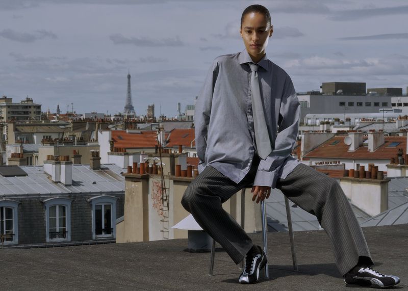 A woman in an oversized shirt and pants, seated on a metal stool, poses on a rooftop with a Paris cityscape and the Eiffel Tower in the background.