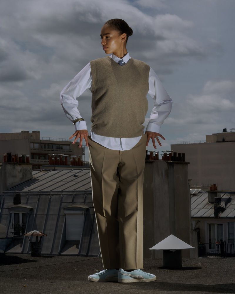 Full-length portrait of a slender person in a sweater vest and trousers standing on a rooftop with buildings and cloudy sky in the background.