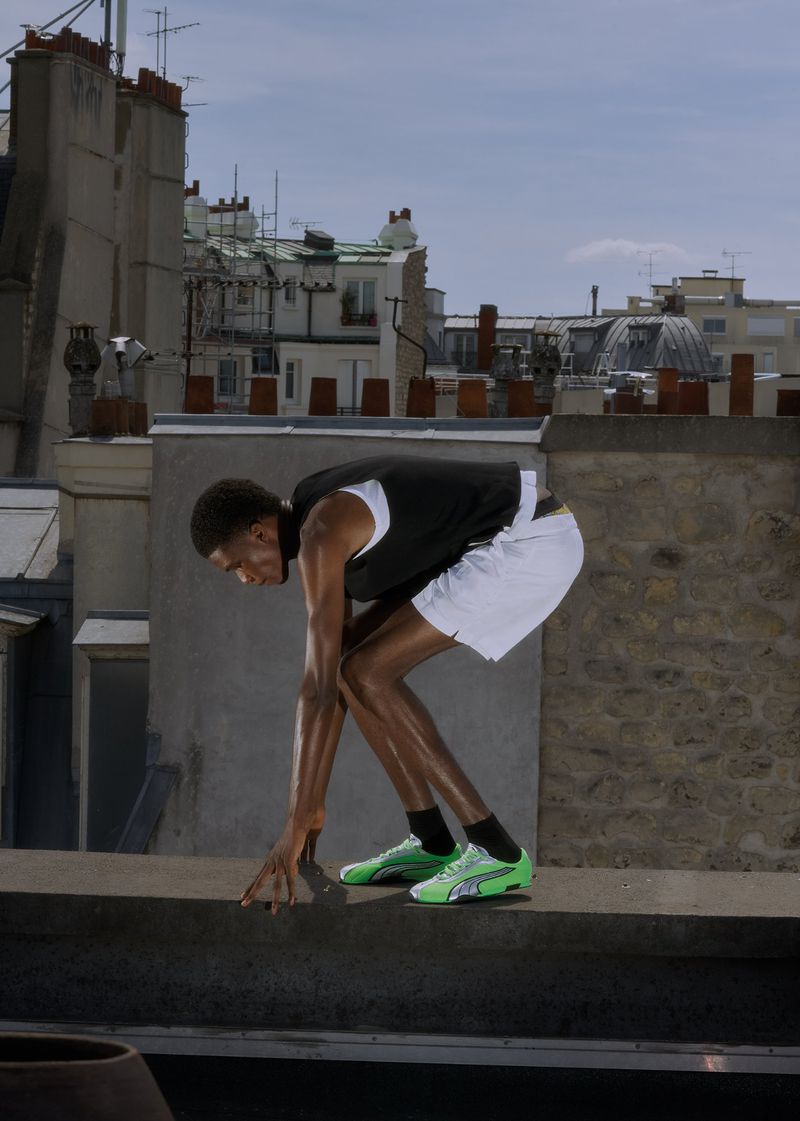 Athletic man in running stance on rooftop with city buildings and blue sky, wearing sport clothes and bright green running shoes.