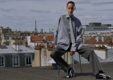 Model in an oversized grey shirt, tie, and pinstripe trousers seated on a stool on a Paris rooftop with the Eiffel Tower in the background.