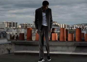 Tall man standing on a city rooftop wearing a grey blazer over a white t-shirt against a cloudy sky