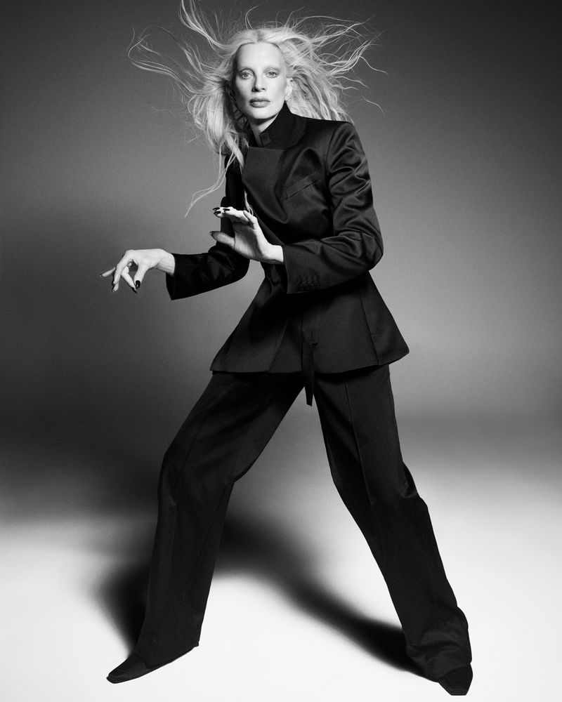 Model Kristen McMenamy poses dynamically in a tailored black suit with windblown platinum hair on a studio set.