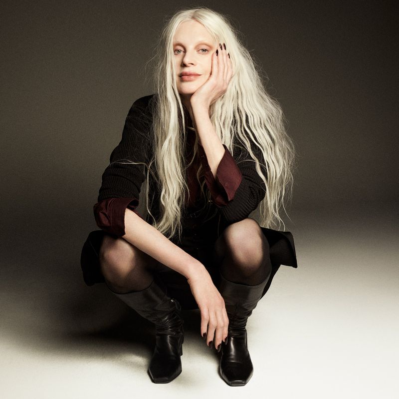 Kristen McMenamy with long white hair squatting in black attire against a grey studio backdrop