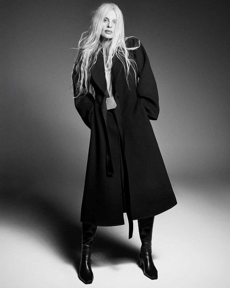 Kristen McMenamy posing in a long black belted coat and leather knee-high boots in a black and white studio portrait.