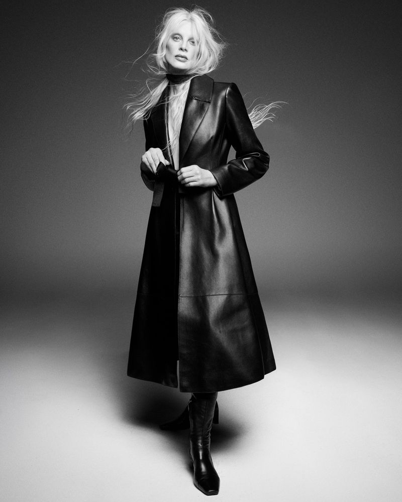 Model Kristen McMenamy posing in a long black leather trench coat and boots in a monochrome studio setting.