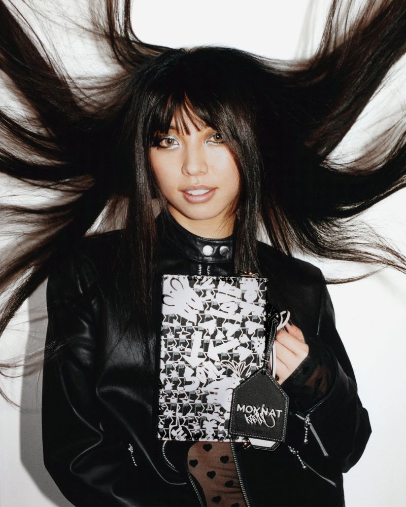 Woman with long dark hair flying outward, wearing a black leather jacket and holding a patterned black and white bag against a white backdrop.