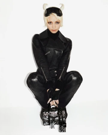 Blonde model in a black leather jumpsuit crouching with a patterned Moynat handbag against a plain white studio background.