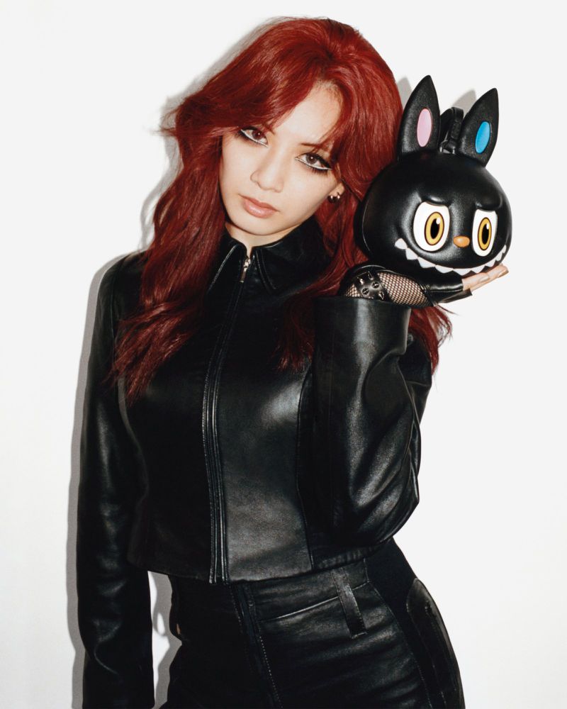 Woman with red hair in a black leather jacket holding a sculpted black character handbag against a white studio wall.