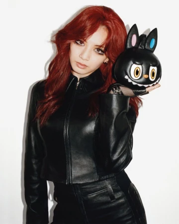 Woman with red hair in a black leather jacket holding a sculpted black character handbag against a white studio wall.