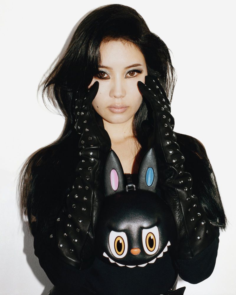 East Asian woman posing with studded black leather gloves and a Moynat x Kasing Lung character bag against a white backdrop.