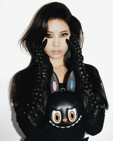 East Asian woman posing with studded black leather gloves and a Moynat x Kasing Lung character bag against a white backdrop.
