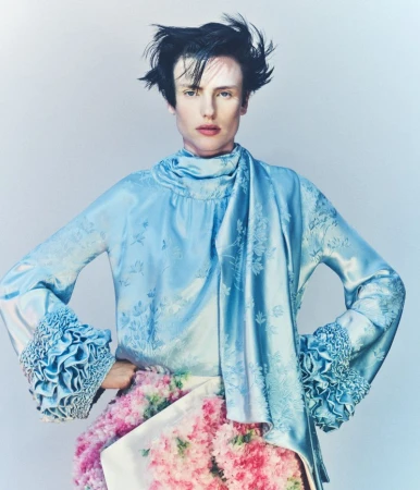 Model Charlie Jones in a light blue silk jacquard top with ruffled cuffs and a pink floral skirt against a pale studio backdrop.