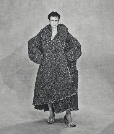 Model Charlie Jones stands wearing a voluminous textured tweed coat with frayed edges and high heels against a mottled grey backdrop.