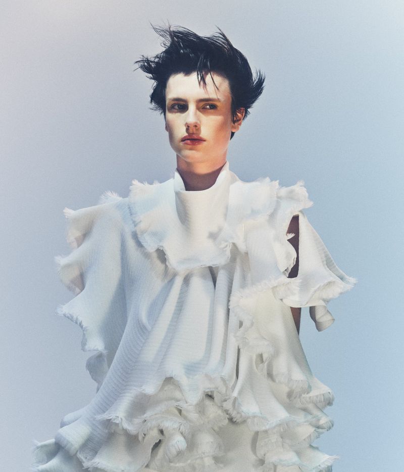 Model Charlie Jones wearing a sculptural ruffled white garment against a soft pale blue gradient backdrop.