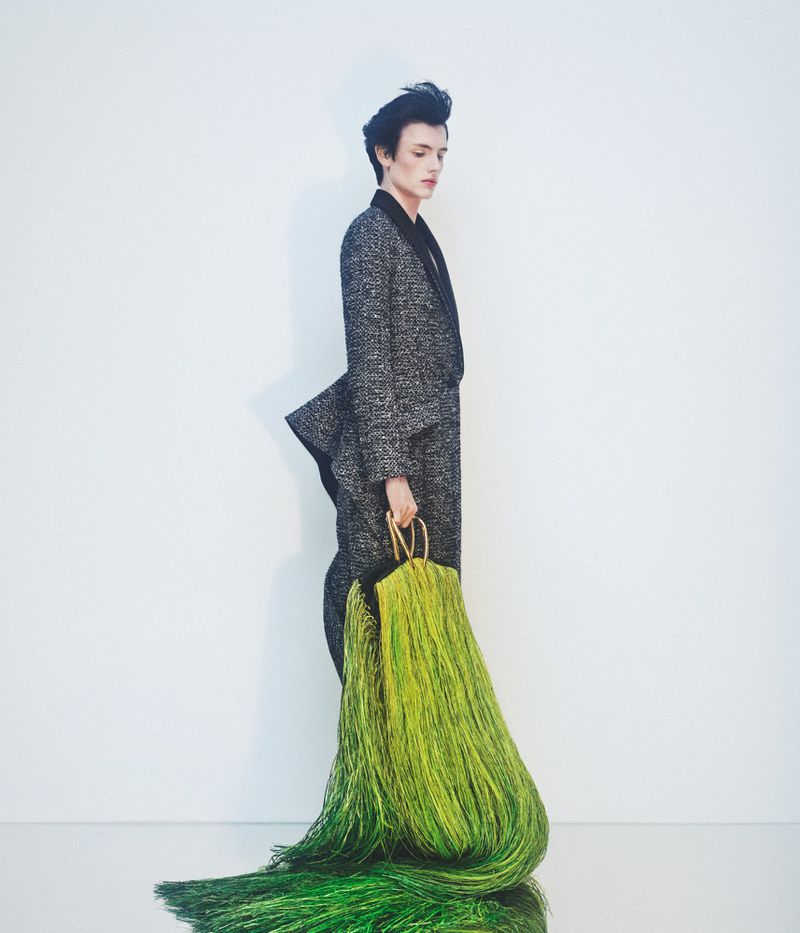 Model in textured dark jacket holding a large flowing green fringed garment against a plain white background