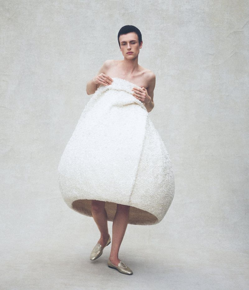Charlie Jones wearing a large sculptural off-white cocoon garment with gold flat shoes against a pale textured studio backdrop.