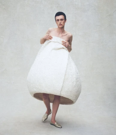 Charlie Jones wearing a large sculptural off-white cocoon garment with gold flat shoes against a pale textured studio backdrop.