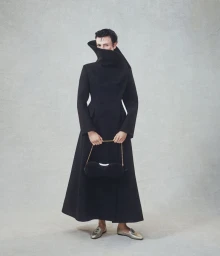 Model Charlie Jones in a high-collared black coat and metallic loafers holding a clutch against a grey studio backdrop.