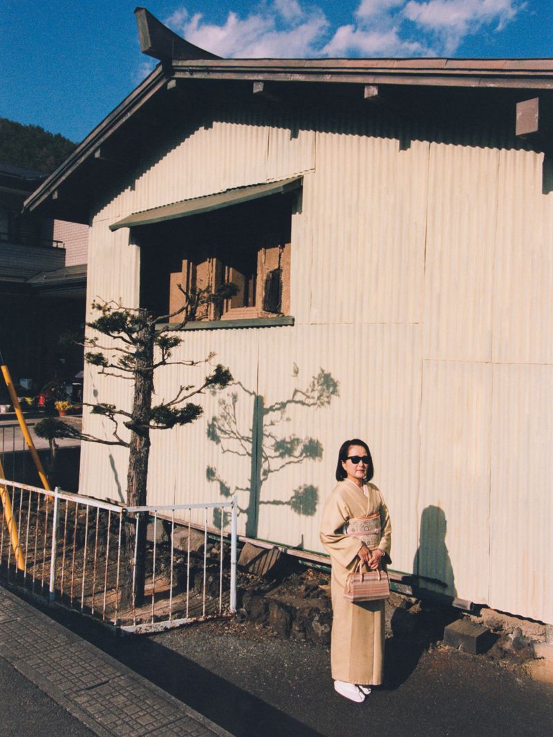 Woman wearing a beige kimono and sunglasses holding a handbag outside in front of a corrugated building wall.