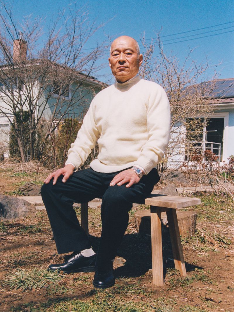 Elderly man in a cream sweater and black trousers seated on a wooden stool outdoors with a house and clear blue sky behind him.