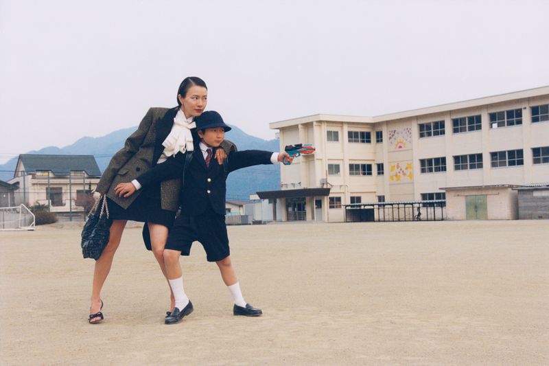 An adult woman and a young boy standing on a school yard pavement in front of a building exterior, with mountains in the background.