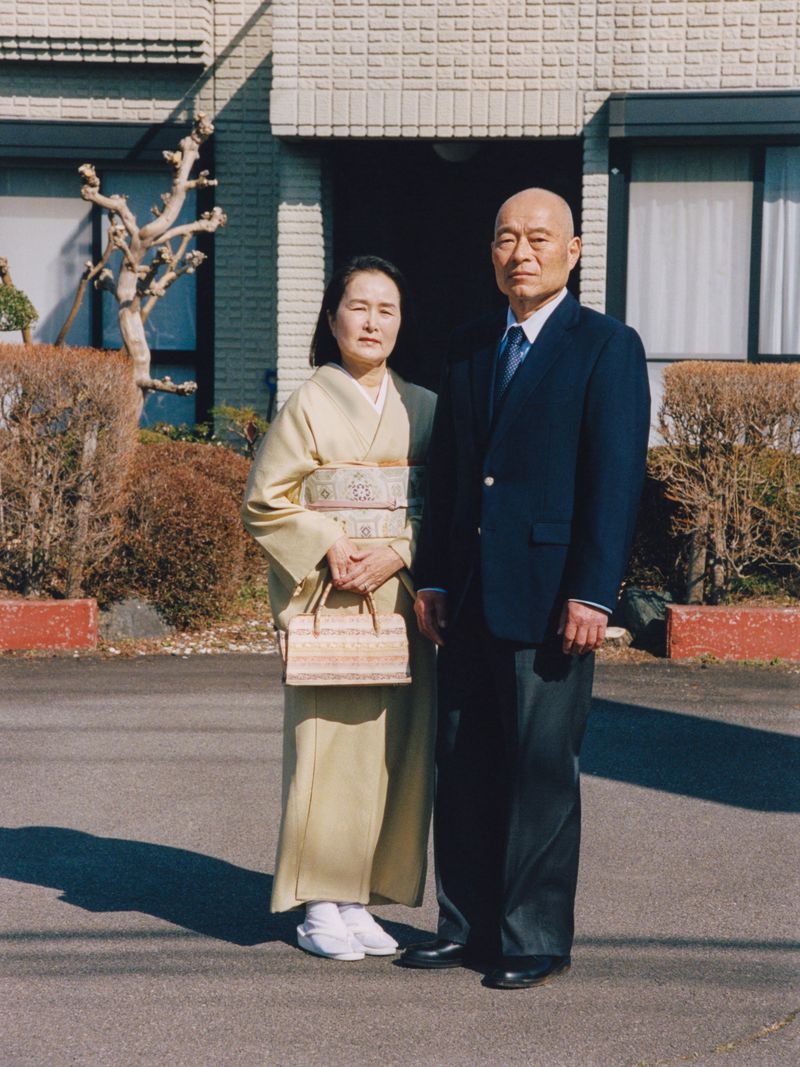Japanese couple standing together outdoors, the woman wearing a cream kimono with a patterned obi and the man in a navy suit jacket.