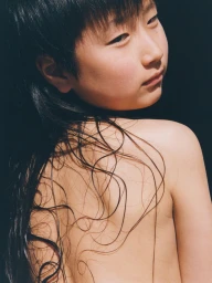Close-up portrait of an East Asian youth with wet black hair strands across a bare back, looking over their shoulder against black.