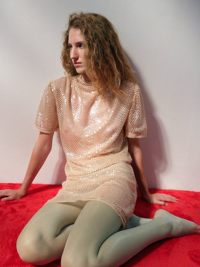 Model Anna Tihonchuk seated on a red plush carpet wearing a sparkling sequin short dress and mint green tights against a white wall.