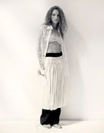 Anna Tihonchuk in a sheer lace shirt and layered white fringe skirt leaning against a studio wall.