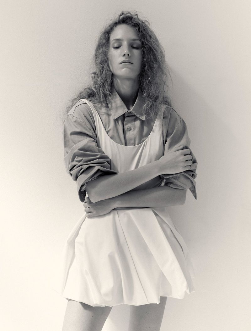 Model Anna Tihonchuk posing with eyes closed in a layered white dress over a collared shirt against a white backdrop.
