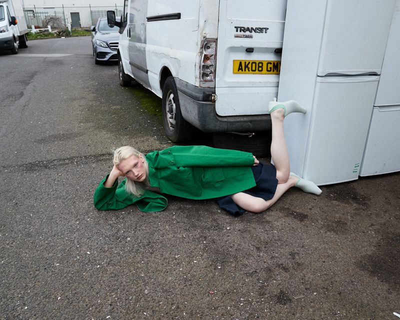 Model lying on asphalt in a green jacket and dark skirt with legs propped against a white refrigerator next to a white van.