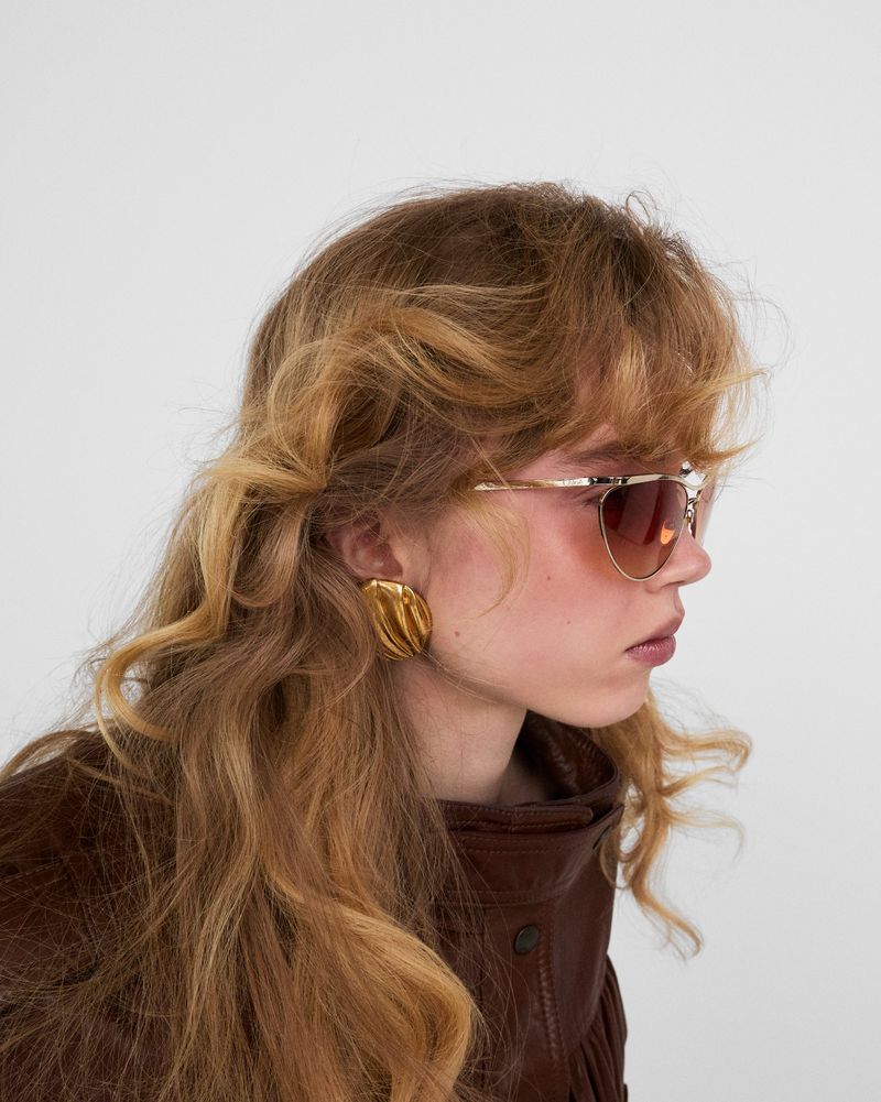 Model in profile wearing tinted aviator sunglasses, a large sculptural gold earring, and a brown leather jacket against a white studio background.