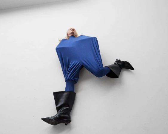 Model lying down wearing an angular cobalt blue stretched jumpsuit and black wide-shaft leather boots on a white surface.