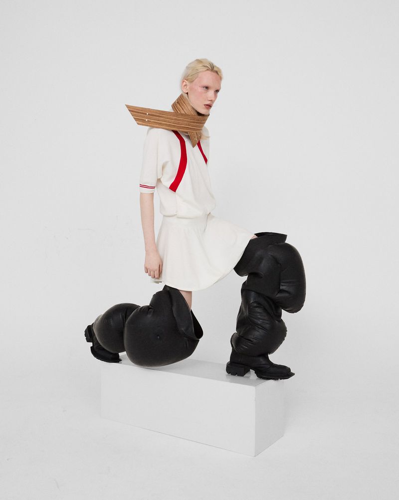 Model posing in white dress, sculptural wooden collar, and exaggerated oversized black puffy legwear on a white studio plinth.