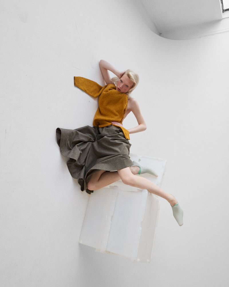 Model posing dynamically in a mustard knit top and pleated grey skirt against a white studio background.