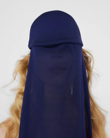 Model with wavy blonde hair wearing a deep navy draped fabric headpiece covering her face against an off-white background.