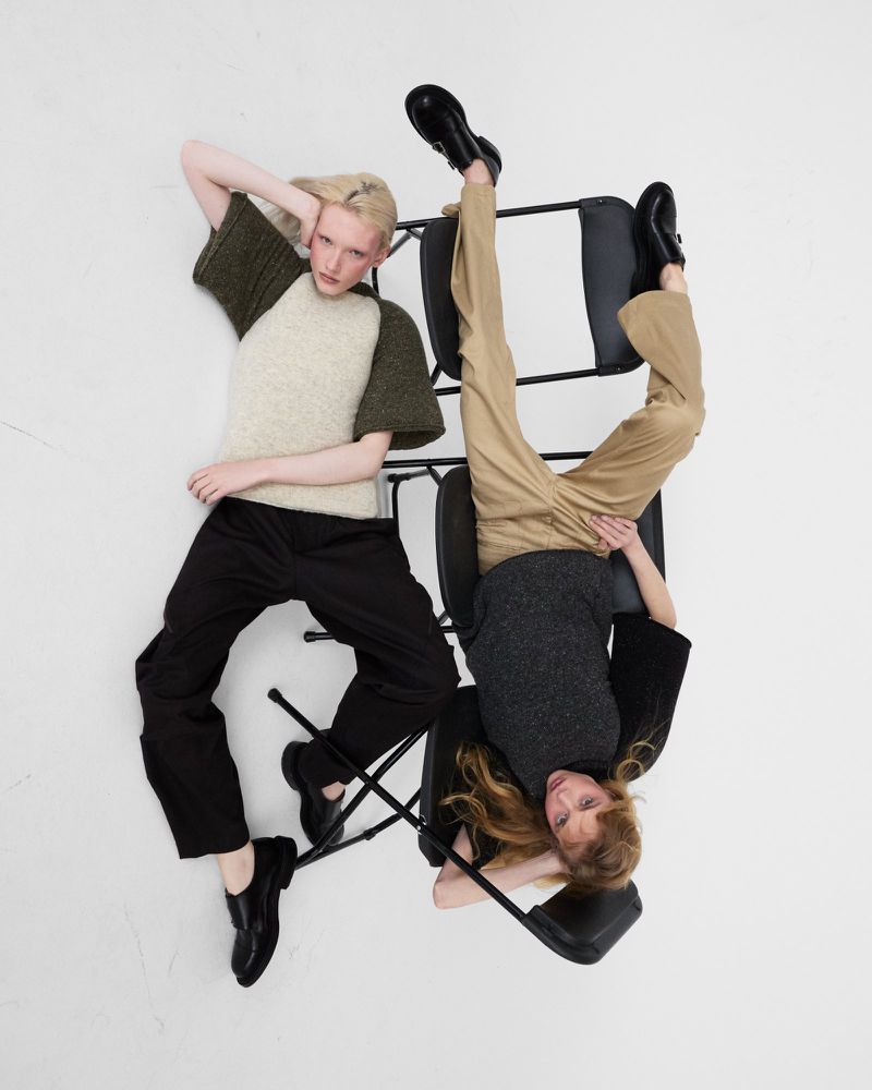 Two fashion models lying head-to-toe across black folding chairs against a clean white seamless studio background.