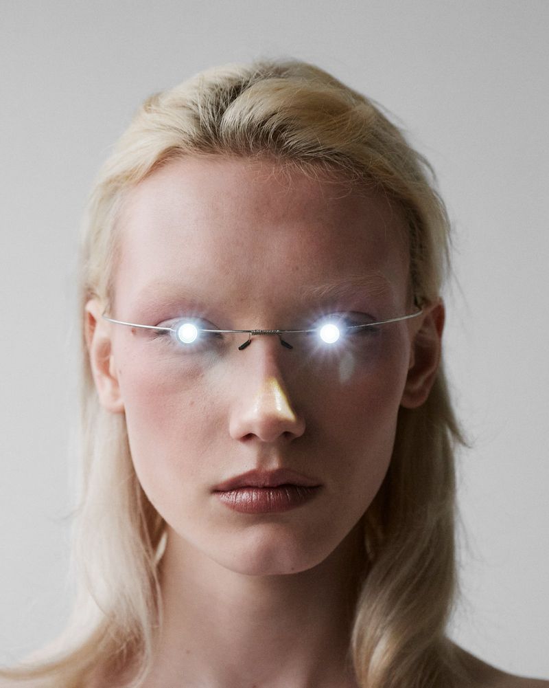 Close-up beauty portrait of a blonde model wearing thin wire glasses with bright glowing circular lights over the eyes.