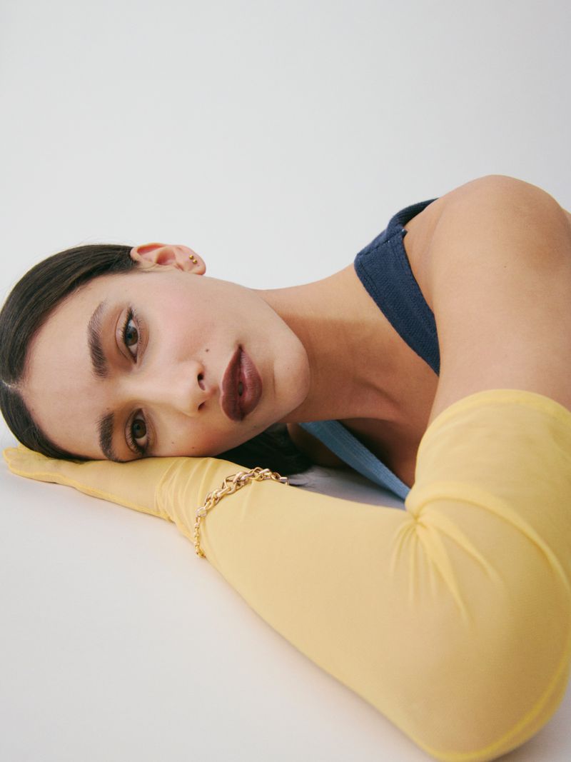 Model Titia Le Roux resting her head on sheer yellow opera gloves wearing dark lipstick against a white studio background.