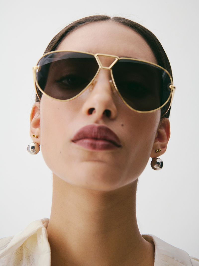 Close-up portrait of a woman wearing gold aviator sunglasses and silver sphere earrings against a plain white background