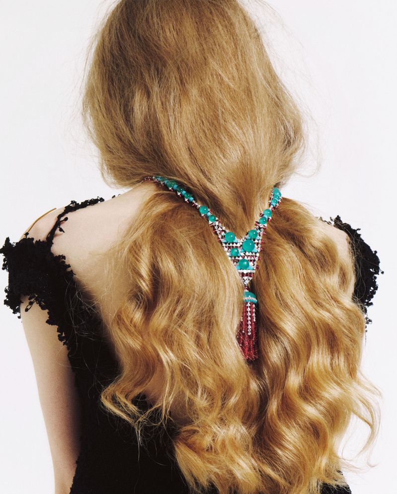 Model with long wavy blonde hair facing away, styled with an ornate turquoise and red tassel necklace, against a white backdrop.