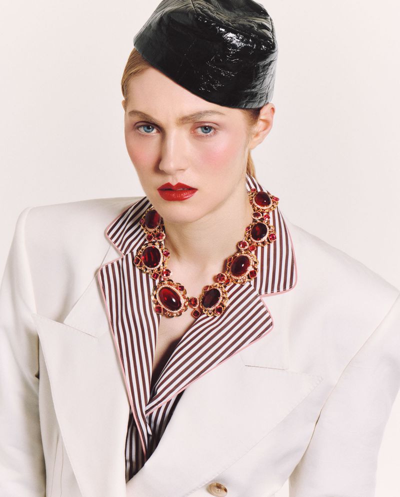 Model wearing a structured white blazer and black cap with statement red gemstone necklace for Vogue Polska