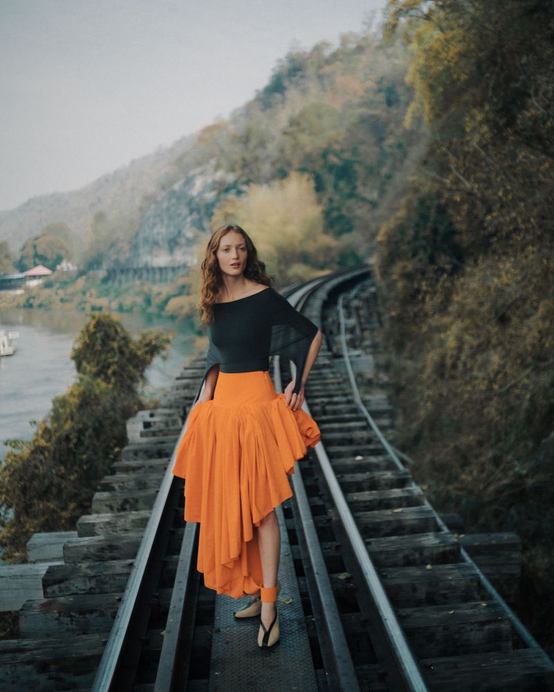 Lydia Reid poses in an orange ruffled skirt and black off-the-shoulder top on elevated railway tracks above a river.