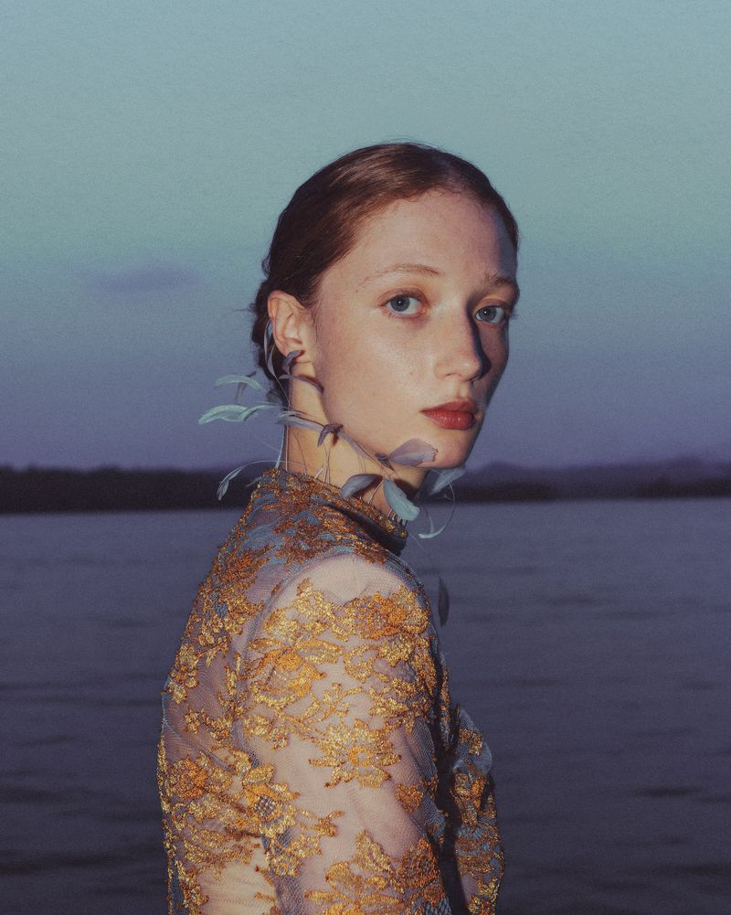 Model Lydia Reid wearing an intricate gold lace top with leaf embellishments by a lake at dusk for Vogue Polska.