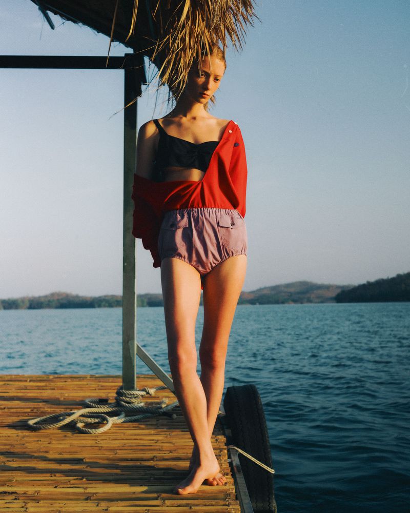 Model standing on a wooden dock by the water wearing a red shirt and high-waisted shorts for Vogue Polska Phuang Malai editorial.